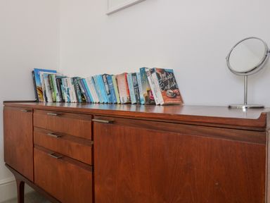 A wooden sideboard with books lined up on top and a small round mirror at The Upper Deck in Porthcurno