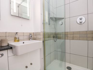 A bathroom with a sink, mirror, soap dispenser, and a glass-enclosed shower at The Upper Deck in Porthcurno