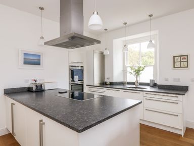 A kitchen with a large island countertop a built-in oven and a window with flowers in a vase at The Upper Deck in Porthcurno
