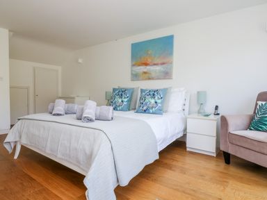 A bedroom with a bed with rolled towels pillows and a painting above it next to a white nightstand and a chair at The Upper Deck in Porthcurno