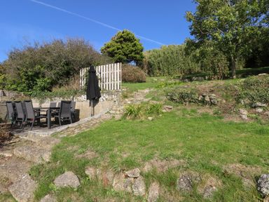 An outdoor stone patio with a dining table and chairs next to a grassy garden with trees and bushes at The Upper Deck in Porthcurno