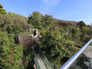 An outdoor area with a glass bridge leading to a patio with chairs and an umbrella surrounded by trees and bushes at The Upper Deck in Porthcurno
