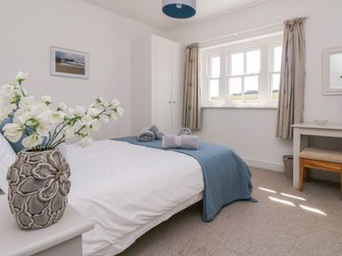 A bedroom with a bed, blue throw, rolled towels, a vase with white flowers, wardrobe, window with curtains, and a small desk at The Upper Deck in Porthcurno