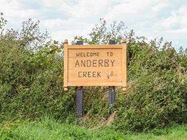 A wooden sign that says welcome to anderby creek in front of bushes and grass