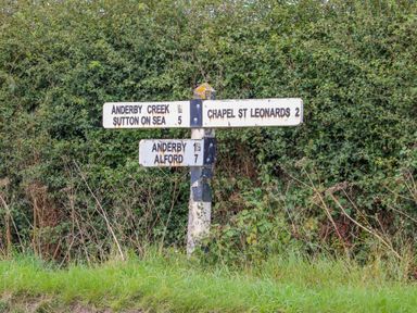 A road sign with directions to Anderby Creek Sutton on Sea Chapel St Leonards Anderby and Alford by a green hedge