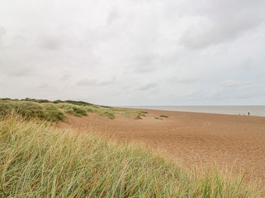 A sandy beach with sparse grass and a person walking near the water at Moggs Retreat in Chapel St Leonards