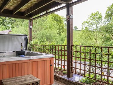 An outdoor hot tub with a bottle of wine and glass on the edge under a wooden roof with a wooden bench beside it overlooking green trees and plants at Valley Lodge 52 in Callington