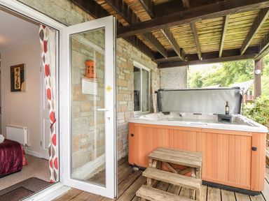 An outdoor covered patio with a wooden hot tub and steps next to a glass door leading to a room with a red bedspread at Valley Lodge 52 Callington