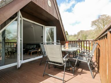 An outdoor wooden deck with a glass table and six gray chairs outside a wooden house with open double doors at Valley Lodge 52 Callington