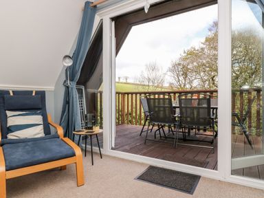 A lounge chair with a side table and a floor lamp next to glass doors leading to a wooden balcony with a table and chairs at Valley Lodge 52 in Callington