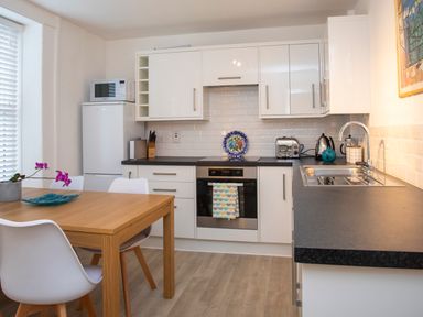 A kitchen with white chairs wooden table white cabinets and black countertop at Bryn Teg Cottage in Menai Bridge