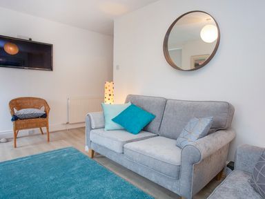 A living room with a grey sofa with cushions a round mirror on the wall a wicker chair with a cushion a mounted TV and a floor lamp at Bryn Teg Cottage in Menai Bridge