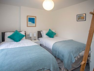 A bedroom with two single beds each with teal pillows and blankets a white nightstand with two lamps and framed pictures on the walls at Bryn Teg Cottage in Menai Bridge