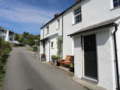 A white exterior of a cottage with a black door and wooden bench on a narrow road at Bryn Teg Cottage in Menai Bridge