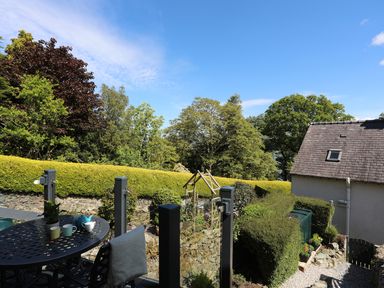 An outdoor patio with a round metal table and chairs overlooking a garden with hedges and trees at Bryn Teg Cottage in Menai Bridge