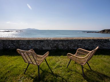 Two outdoor wicker chairs on grass facing a stone wall with a view of the sea and distant hills at Stabl Bach in Llanfaethlu