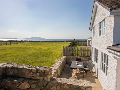 A patio with a table and chairs beside a white house overlooking a fenced grassy area and the sea at Stabl Bach in Llanfaethlu