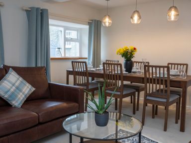 A dining room with a brown leather sofa a glass coffee table with a plant and a wooden dining table set with flowers at Stabl Bach in Llanfaethlu