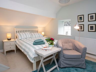 A bedroom with a bed striped bedding two nightstands lamps a gray armchair a small table with books and a plant a window and framed pictures on the wall at Stabl Bach in Llanfaethlu