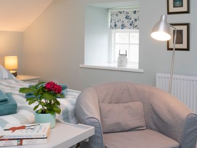 A bedroom with a grey armchair beside a small table with books and a plant at Stabl Bach in Llanfaethlu