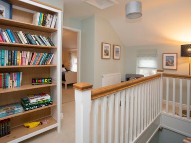 A hallway with a wooden bookcase filled with books and board games next to a staircase with white banisters at Stabl Bach in Llanfaethlu