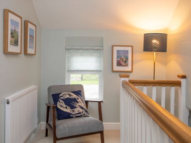 A small sitting area with a gray chair and blue lobster pillow near a window and a floor lamp at Stabl Bach in Llanfaethlu