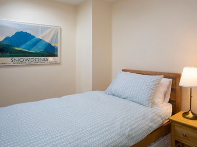 A bedroom with a bed with patterned bedding a wooden bedside table with a lamp and a framed mountain poster on the wall at Stabl Bach in Llanfaethlu