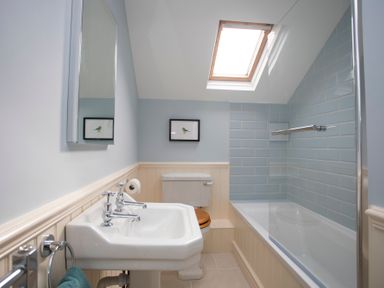 A bathroom with a sink, toilet with wooden seat, bathtub with glass screen under a skylight at Stabl Bach in Llanfaethlu