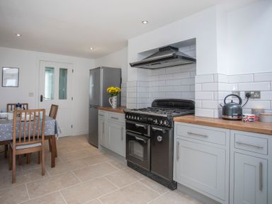 A kitchen with a black stove, grey cabinets, a silver refrigerator, a dining table with chairs, and a kettle on the counter at Stabl Bach in Llanfaethlu
