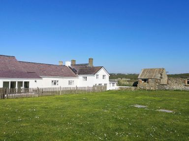 A white house with multiple chimneys and a wooden fence on a grassy field with a small stone building at Stabl Bach in Llanfaethlu