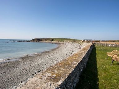 A pebble beach with a stone wall and two wicker chairs on grass at Stabl Bach in Llanfaethlu