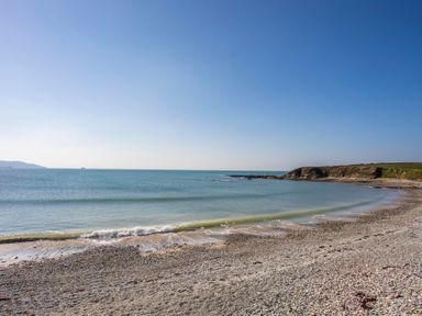 A pebble beach with gentle waves and a rocky headland under a clear sky at Stabl Bach in Llanfaethlu