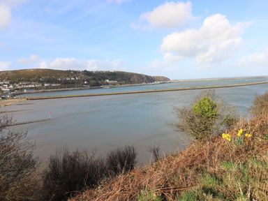 A river with a rocky breakwater and buildings on a hillside under a blue sky with clouds