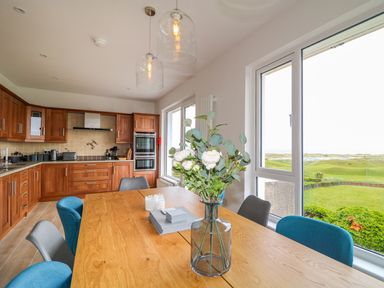 A kitchen with wooden cabinets a dining table with chairs and a vase with flowers by large windows showing a green landscape at Seacoast Cottage in Castlerock