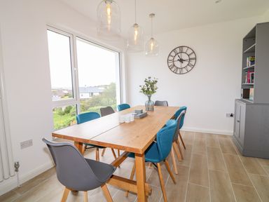 A dining area with a wooden table and blue and gray chairs a clock on the wall and a cabinet with books at Seacoast Cottage in Castlerock