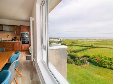 A kitchen with wooden cabinets and blue chairs next to a window showing a green landscape and ocean view at Seacoast Cottage in Castlerock