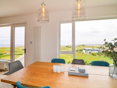 A dining area with a wooden table and chairs near large windows overlooking green fields and distant buildings at Seacoast Cottage in Castlerock