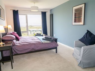 A bedroom with a bed and nightstand with lamps next to a window and a gray armchair with a pillow at Seacoast Cottage in Castlerock