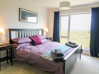 A bedroom with a black bed frame purple bedding folded towels on bed two bedside tables with lamps and large window with blue curtains at Seacoast Cottage in Castlerock