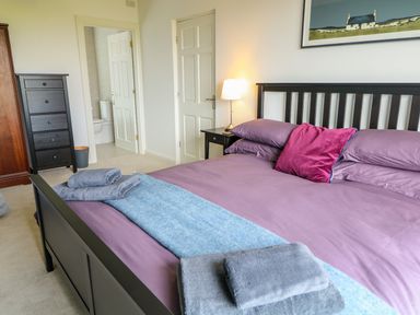 A bedroom with a black bed frame purple bedding gray towels on the bed a nightstand with a lamp and an open door leading to a bathroom at Seacoast Cottage in Castlerock
