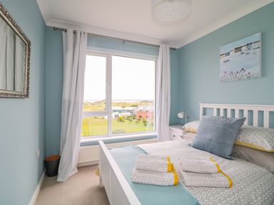 A bedroom with a bed linens and folded towels on it a nightstand with a lamp a large window with white curtains and a framed mirror on the wall at Seacoast Cottage in Castlerock