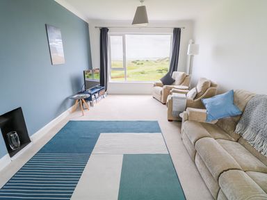 A living room with beige sofa and chairs a blue and white rug a television near a large window with a view of green hills at Seacoast Cottage in Castlerock