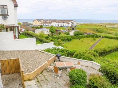 A patio with table and chairs overlooking green lawns and a golf club building by the sea at Seacoast Cottage in Castlerock