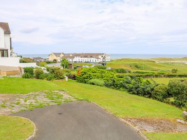 A grassy garden with bushes and a paved path overlooking buildings and the ocean at Seacoast Cottage in Castlerock