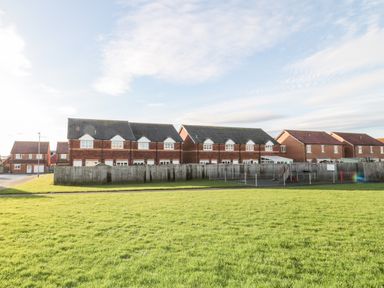 A row of brick houses with tiled roofs behind a fenced playground and green grass in front at 39 Longbeach Drive in Beadnell