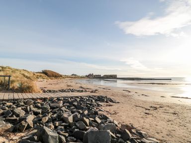 A sandy beach with rocks in the foreground grassy dunes on the left and buildings in the distance at 39 Longbeach Drive in Beadnell