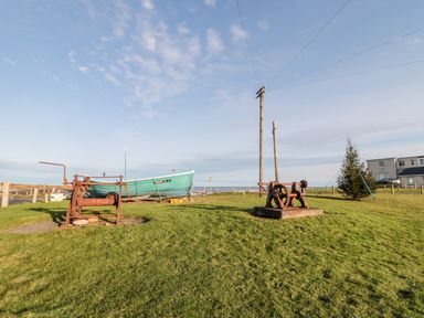 A grassy area with two rusty winches a fishing boat and a house near the sea at 39 Longbeach Drive in Beadnell
