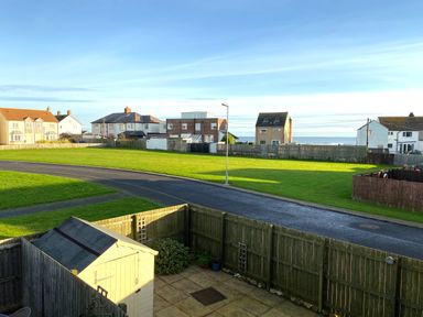 A fenced backyard with a small shed and plants overlooking a road and green grassy area with houses and the sea in the background at 39 Longbeach Drive Beadnell