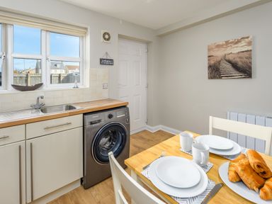 A kitchen with a washing machine under a wooden countertop a dining table set with plates mugs and pastries on a plate at 39 Longbeach Drive in Beadnell