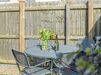 An outdoor metal table with four matching chairs a vase with flowers two wine glasses and a bottle of wine on the table near a wooden fence 39 Longbeach Drive Beadnell